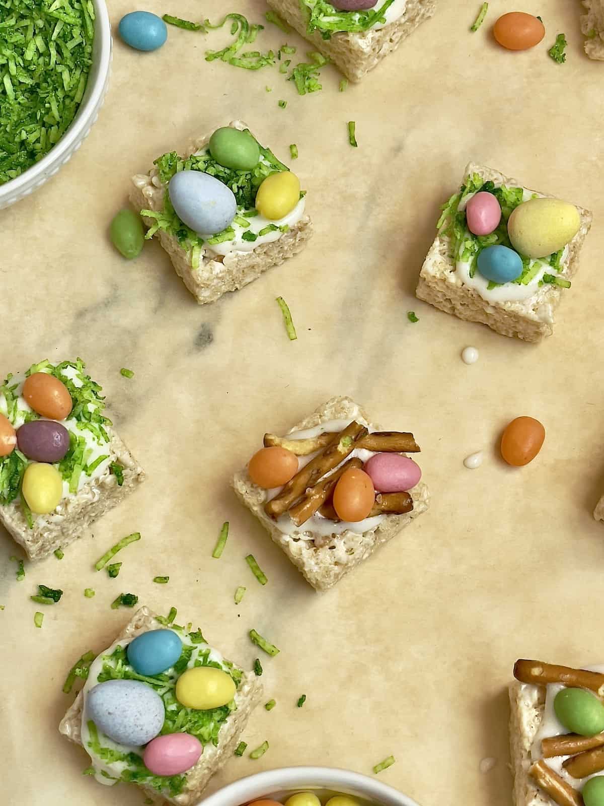 mini egg rice krispie squares on parchment paper decorated with coconut grass, jelly beans, and pretzel sticks