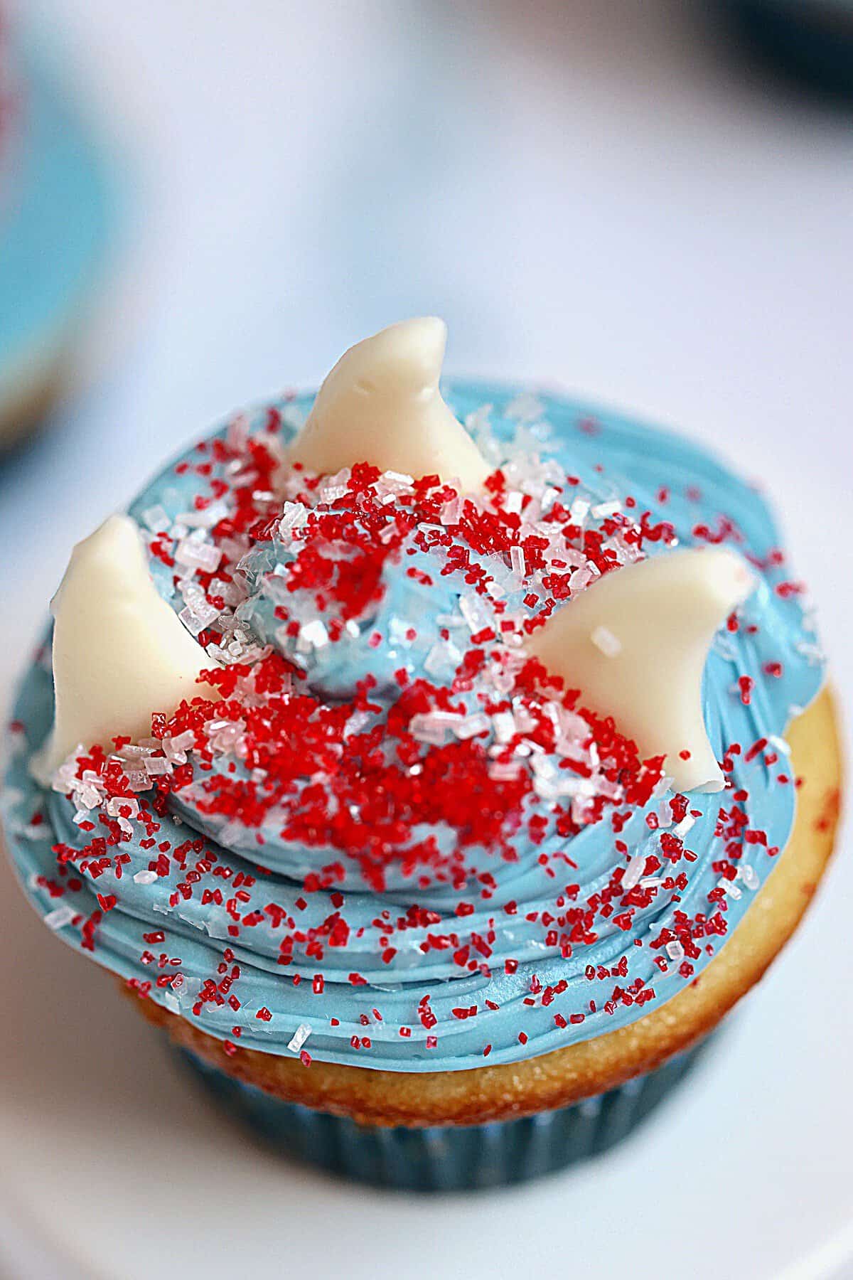 shark cupcake with 3 white shark fins on blue frosting and decorated with red and white sprinkles