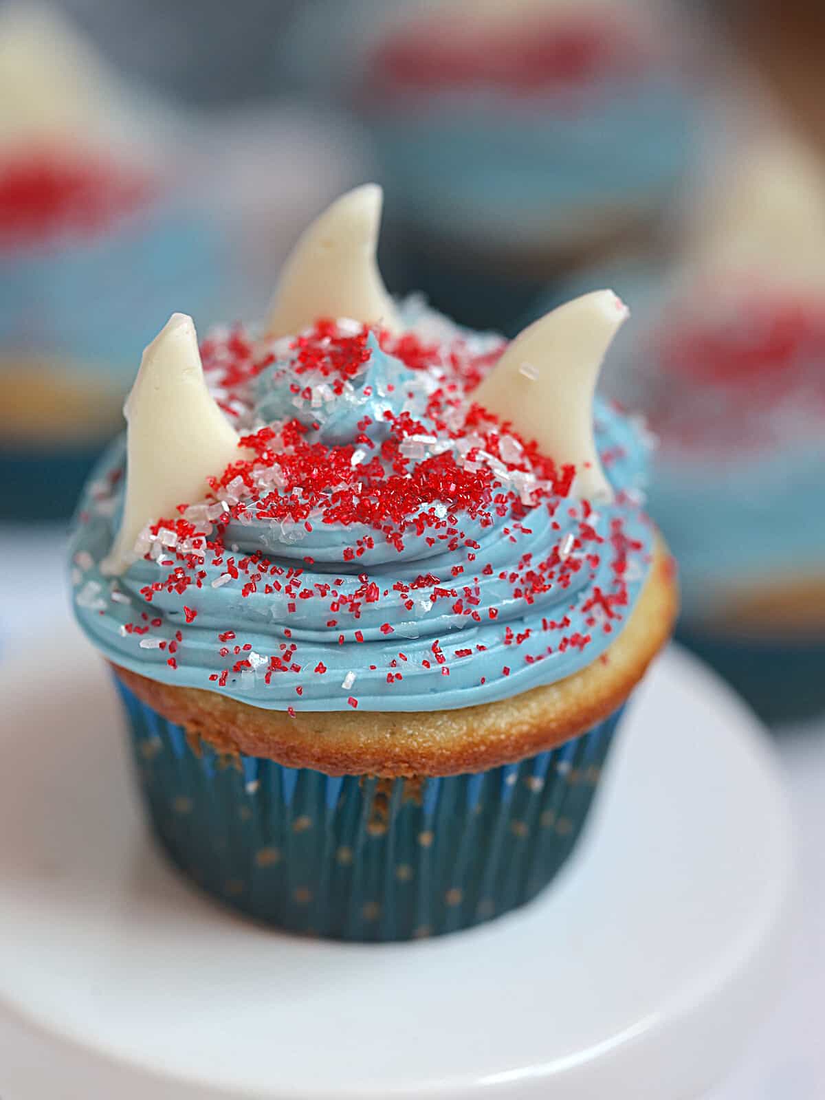 blue cupcakes with blue frosting, red and white sprinkles and white shark fins on a white cupcake stand