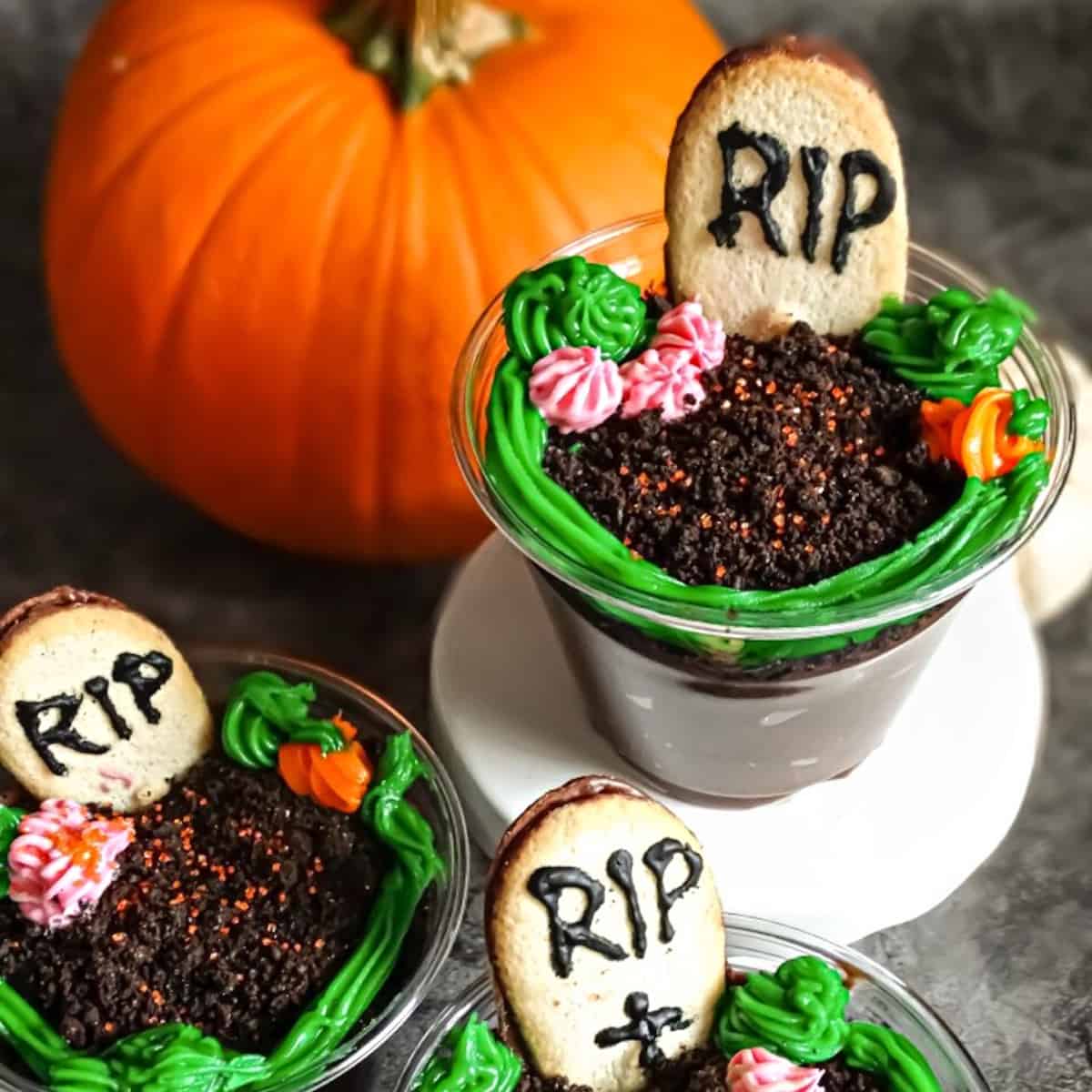 three chocolate graveyard pudding cups decorated with a Milano cookie headstone and pumpkin and flowers