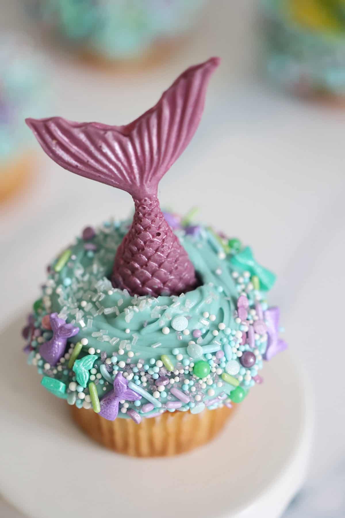 mermaid cupcake with a purple tail blue icing and mermaid sprinkles on a cake stand