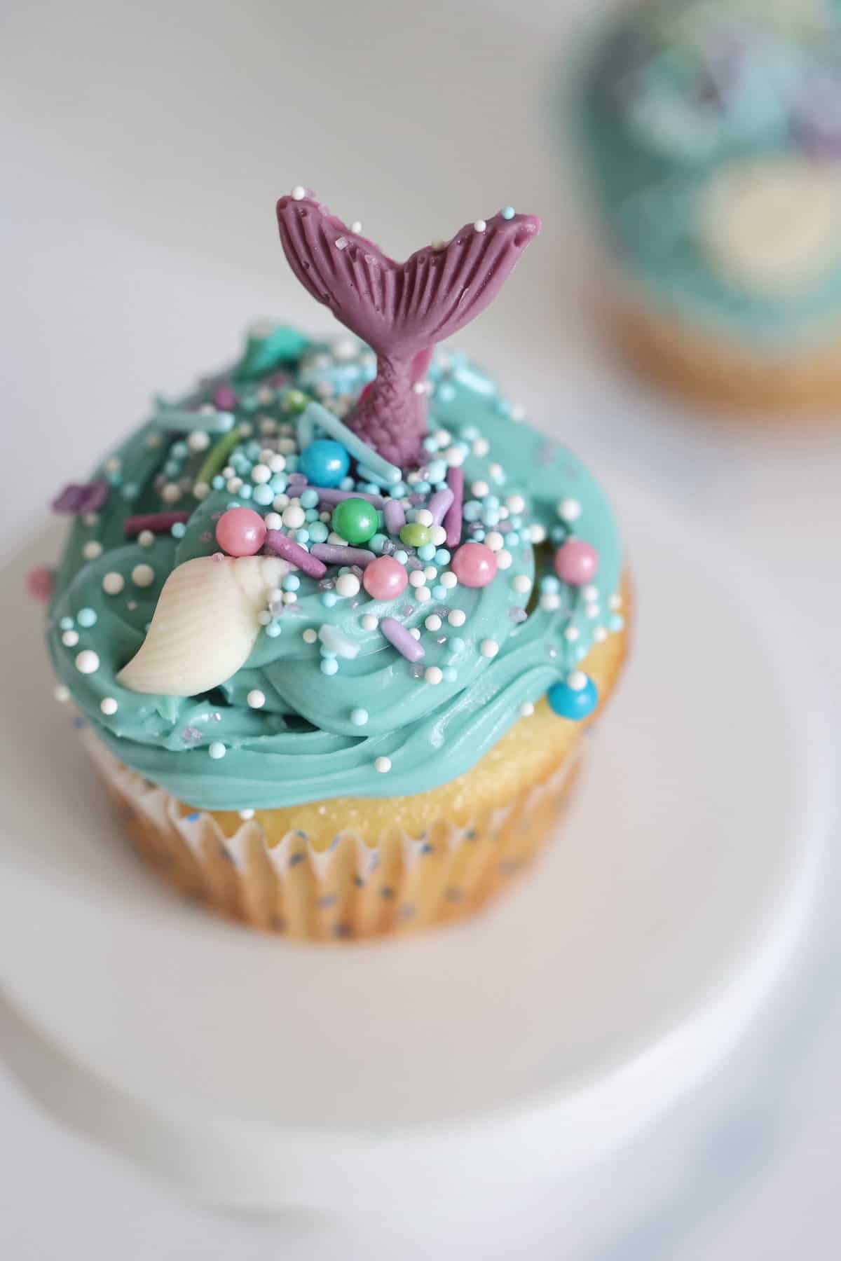mermaid cupcake with a purple tail blue icing and mermaid sprinkles on a cake stand