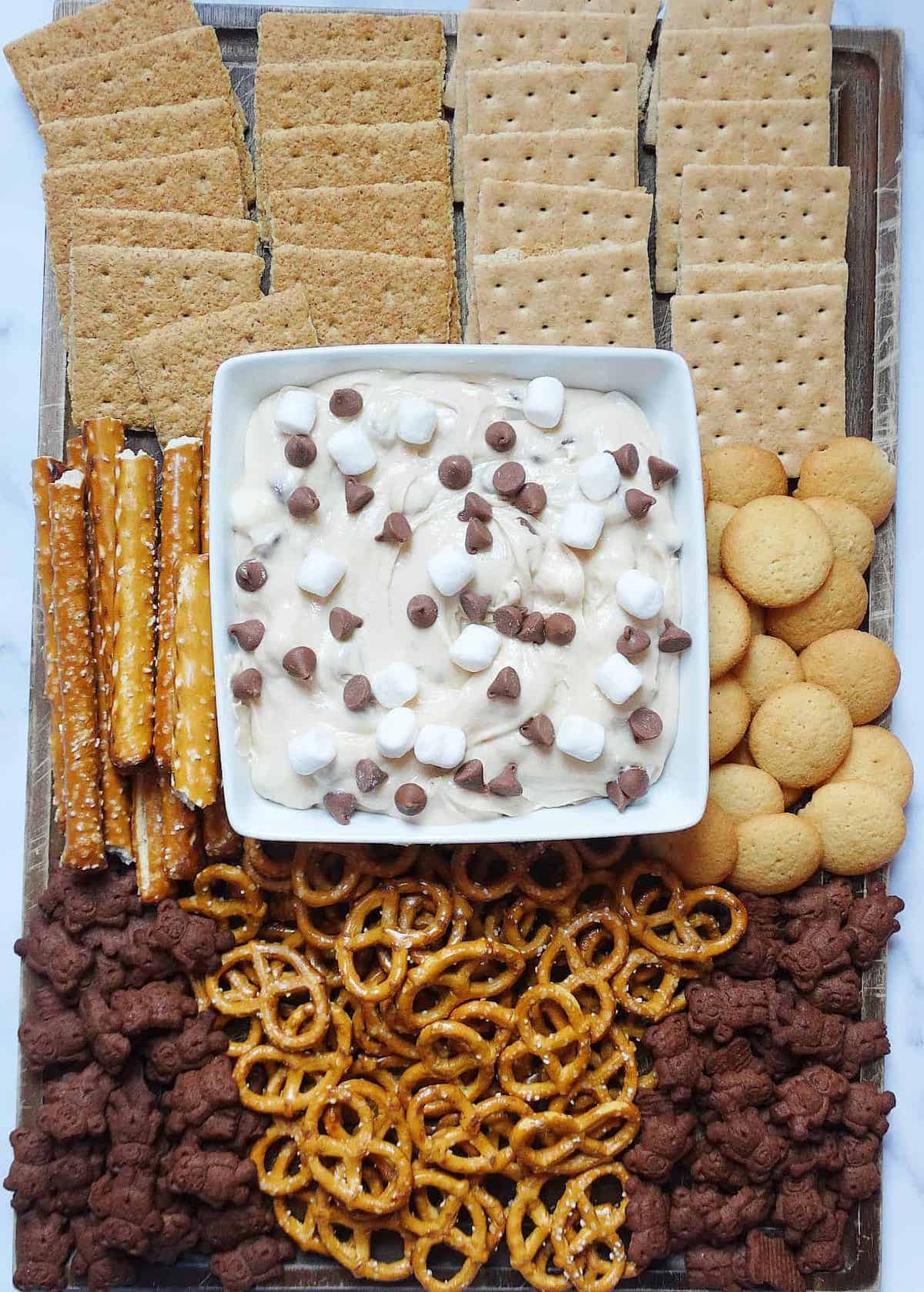 s'mores dip topped with chocolate chips and marshmallows in a white serving bowl on a charcuterie board with crackers