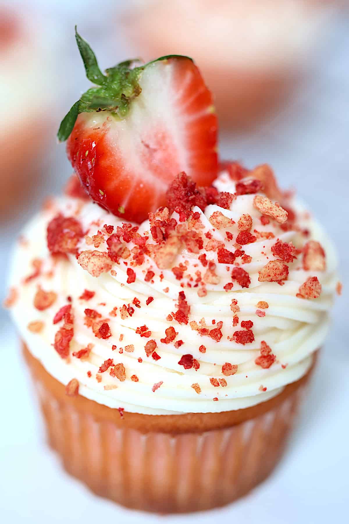 strawberry crunch cupcake on a white cake plate frosted with buttercream and topped with fresh strawberry and crunchy topping