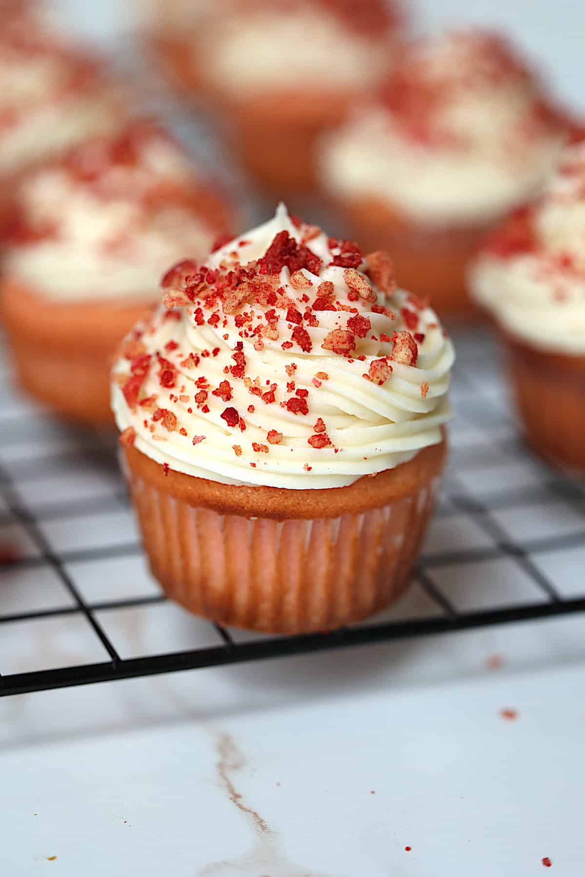 a strawberry cupcake topped with frosting and strawberry crunch on a cooling rack